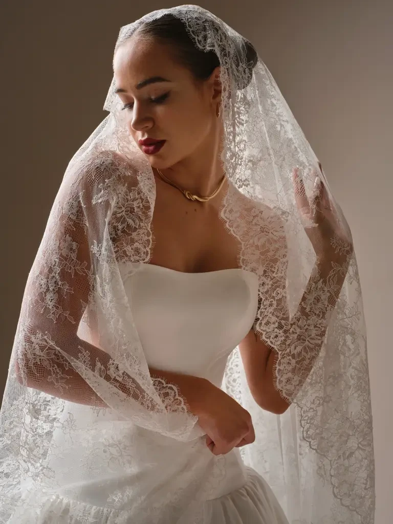 A woman wearing a wedding dress and a lace veil, posing gracefully with her eyes closed.