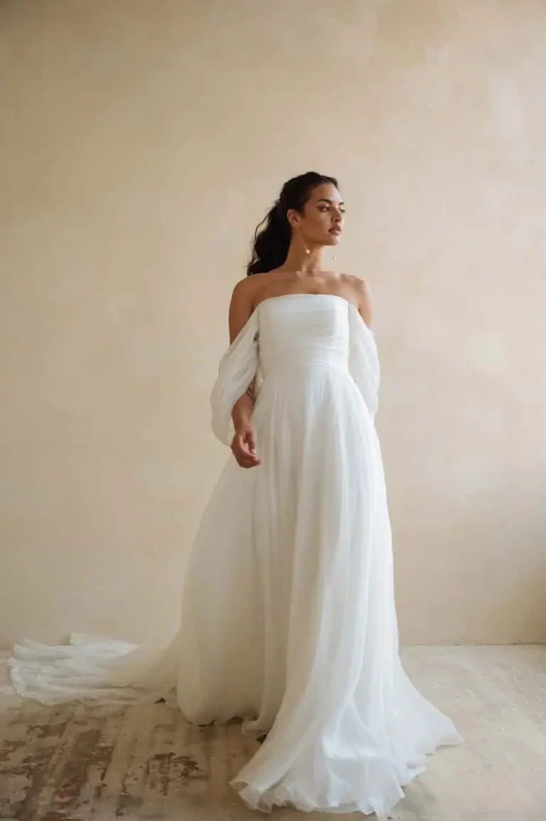 model is wearing a boho chic minimalist a-line wedding dress with off the shoulder sleeves by jenny yoo