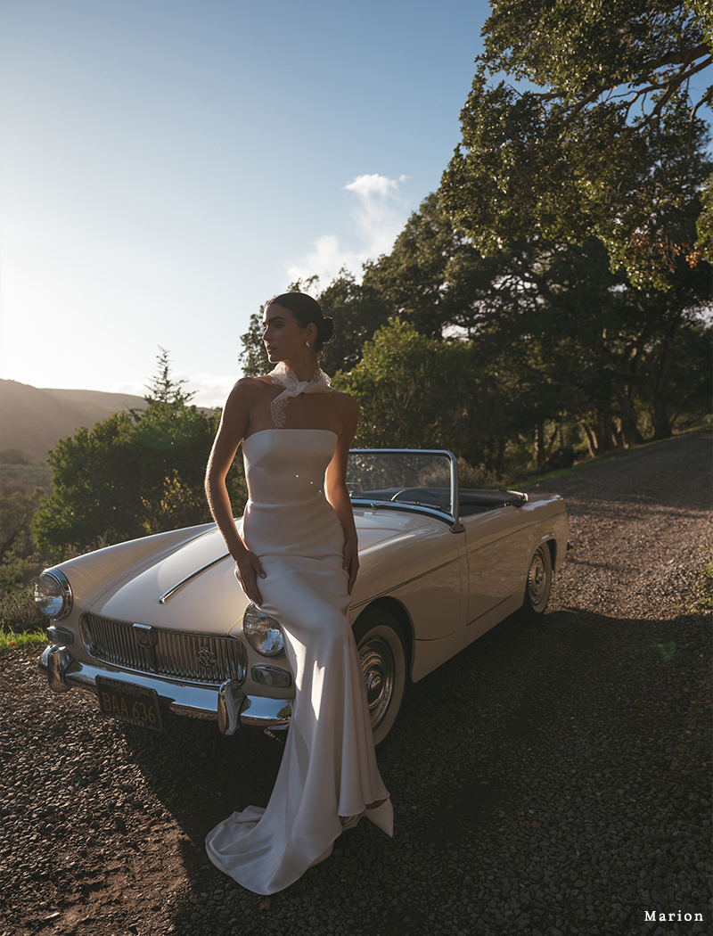 A woman in an elegant white dress leans against a classic convertible on a scenic road. The setting sun casts a warm glow, surrounded by lush greenery.