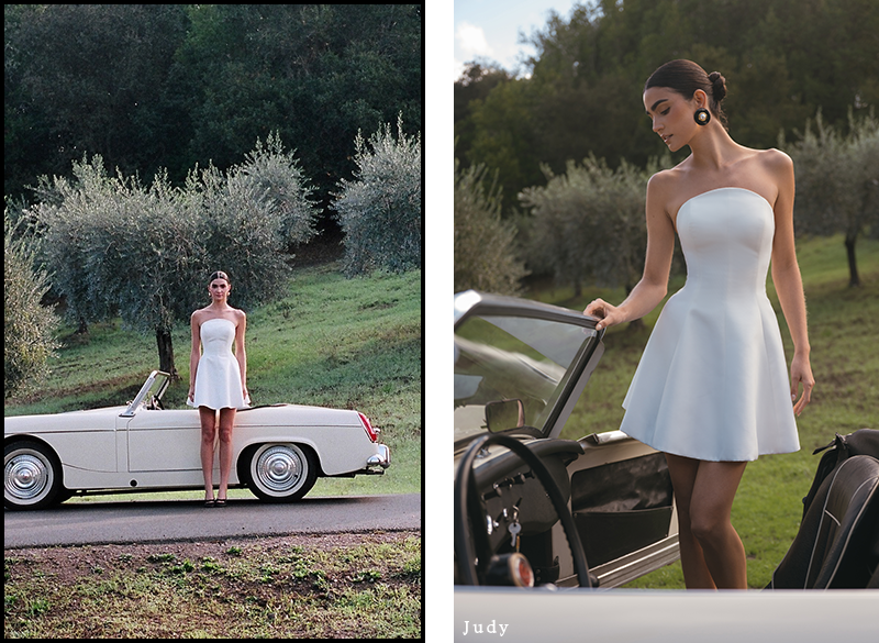 A woman in a white strapless dress stands beside a classic white convertible in a serene, green landscape with olive trees, exuding elegance and tranquility.