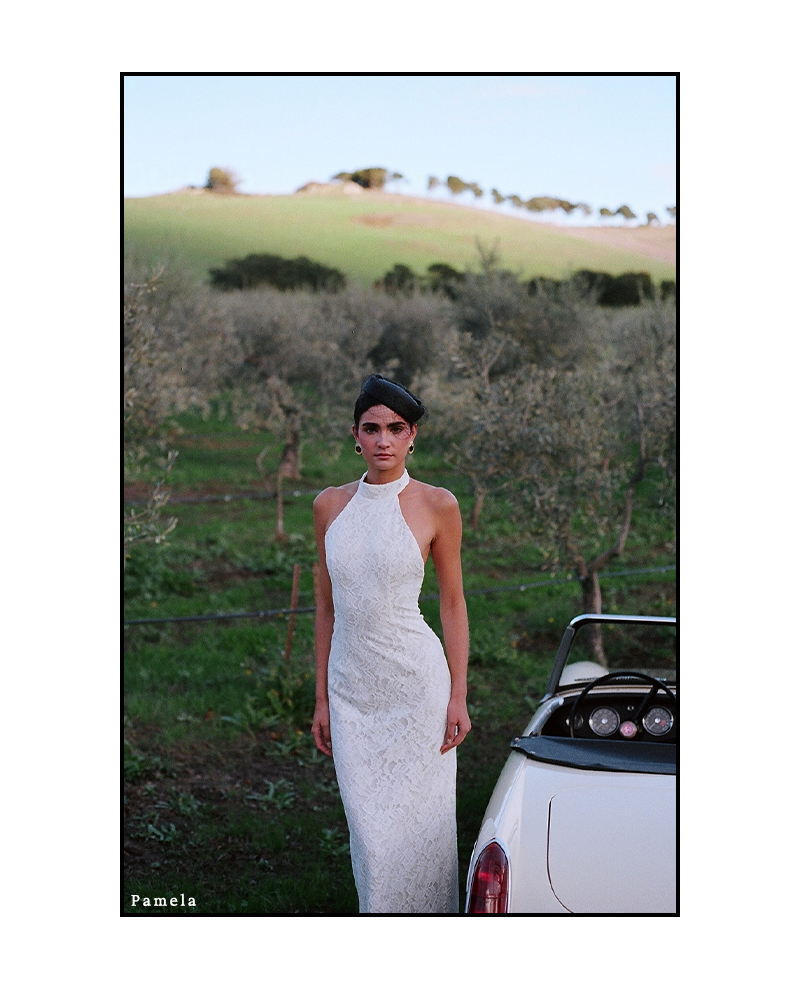 Woman in a white halter dress stands by a vintage convertible in a lush, hilly vineyard. The scene conveys elegance and tranquility.